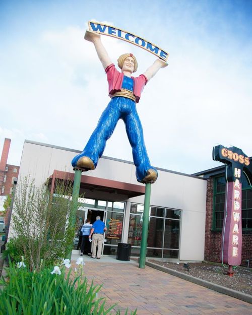 A large statue of a welcoming figure stands outside a hardware store, with people entering the building.