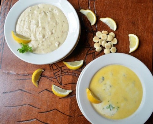 Two bowls of soup on a wooden table, one creamy and the other yellow, garnished with lemon wedges and crackers.