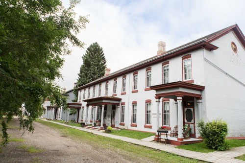 A row of historic buildings with white walls and red trim, surrounded by greenery and a gravel path.