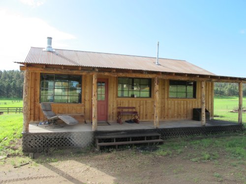 A rustic wooden cabin with a porch, surrounded by green fields and trees under a clear blue sky.
