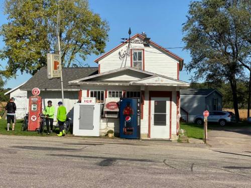 A small, vintage gas station with a Pepsi vending machine and cyclists nearby on a sunny day.