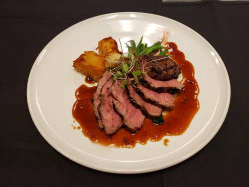 A plate of sliced beef with a rich sauce, garnished with microgreens and accompanied by roasted potatoes.