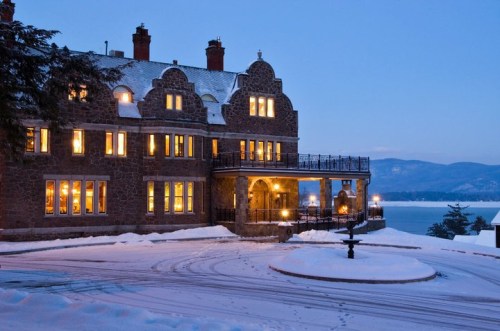 A stone mansion with glowing windows, surrounded by snow, overlooking a lake at dusk.