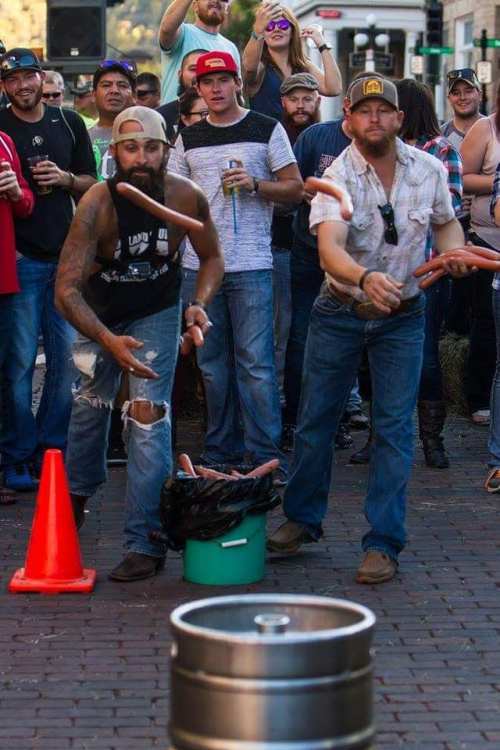 Two men throw objects towards a barrel while a crowd watches, enjoying a lively outdoor event.