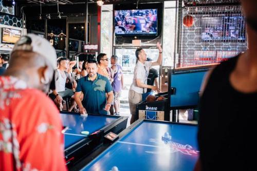 A lively bar scene with people playing air hockey and watching sports on TV in the background.