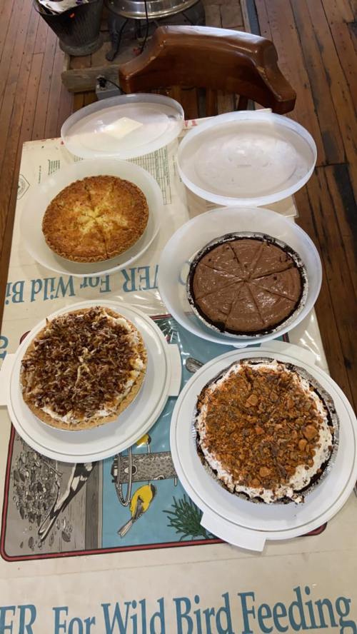 A table displays five assorted pies, including chocolate, coconut, and pecan, in clear containers.