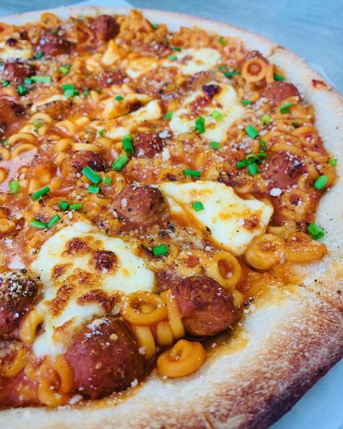 A close-up of a pizza topped with pasta, sausage, cheese, and green onions on a golden crust.