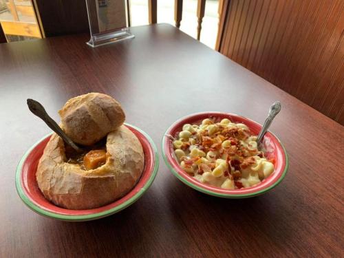 A bread bowl filled with soup next to a bowl of macaroni and cheese topped with bacon bits, on a wooden table.