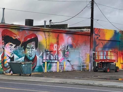 Colorful mural featuring two faces, vibrant graffiti, and a construction lift in front of a cityscape backdrop.