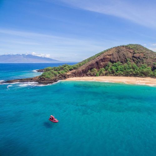 Aerial view of a serene beach with a small red boat near a lush green hill and clear turquoise waters.