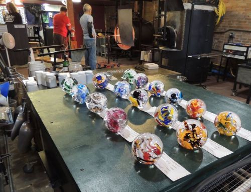 A workshop with colorful glass orbs displayed on a table, while people work in the background.