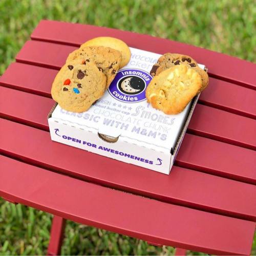 A box of Insomnia Cookies on a red table, featuring various cookies including chocolate chip and s'mores.