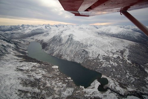 Aerial view of snow-covered mountains and a winding lake under a cloudy sky, seen from a small aircraft.