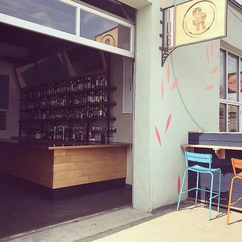 A bar entrance with a large open window, showcasing a wooden bar and colorful outdoor seating.