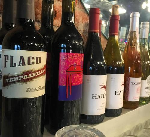 A selection of wine bottles on a shelf, featuring various labels and colors, against a brick wall backdrop.