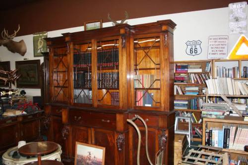 A wooden bookshelf with glass doors, filled with books, in a cozy room with various decor and a rustic atmosphere.