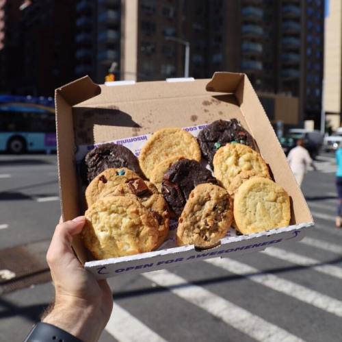A hand holds a box of assorted cookies, including chocolate and vanilla, in a city street setting.