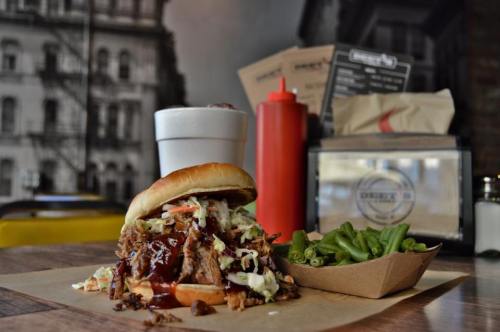 A pulled pork sandwich with coleslaw and BBQ sauce, served with green beans and a drink in a casual dining setting.