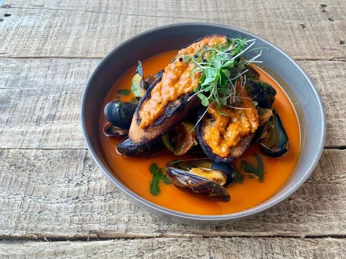 A bowl of mussels in a rich orange sauce, topped with two toasted bread slices and microgreens, on a wooden table.