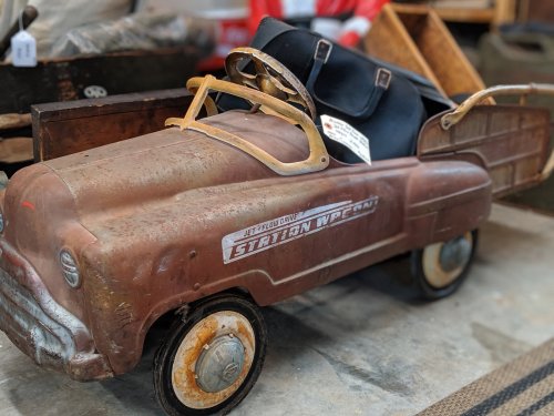A vintage metal toy car with a rusty finish, featuring a black seat and a sign on the dashboard, displayed indoors.