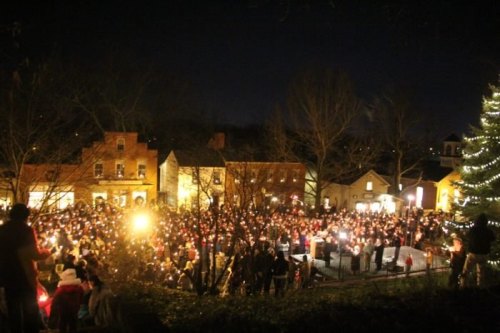 A large crowd gathers at night, holding candles near a decorated tree in a historic village setting.