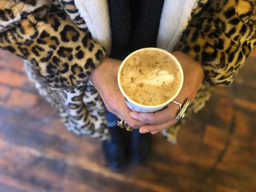 A person in a leopard print coat holds a coffee cup with foam art, surrounded by wooden flooring.