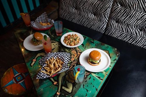 A vibrant table with burgers, fries, and colorful drinks, set against a funky, artistic background.