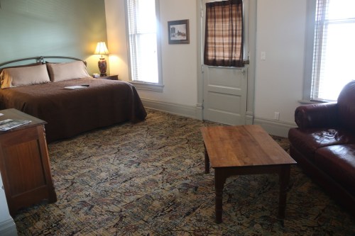 Cozy room with a bed, leather sofa, wooden table, and large windows, featuring warm lighting and patterned carpet.