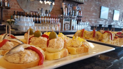 A platter of assorted tapas featuring bread topped with various spreads, garnished with olives and peppers, in a bar setting.