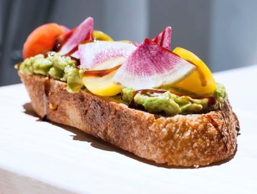 A slice of toasted bread topped with avocado, colorful radishes, and cherry tomatoes, drizzled with balsamic glaze.