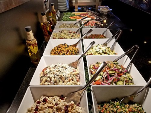 A variety of colorful salads and dishes in white bowls on a buffet table, with olive oil bottles in the background.