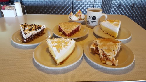 A variety of pies on plates, including chocolate, cherry, lemon meringue, and apple, with a coffee mug in the background.