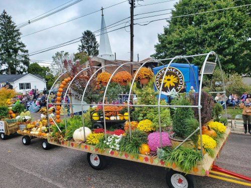 A colorful parade float adorned with pumpkins, flowers, and autumn decorations, passing by a crowd and church steeple.