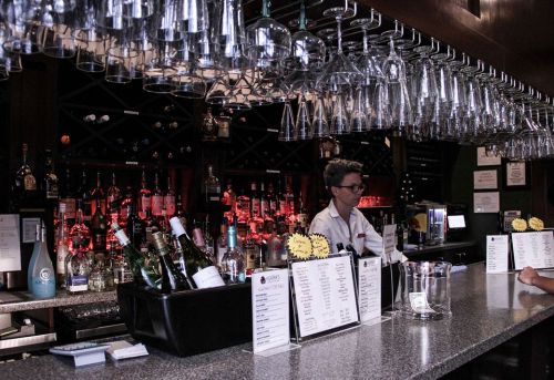 A bartender stands behind a bar filled with bottles and hanging glasses, with a menu displayed in front.