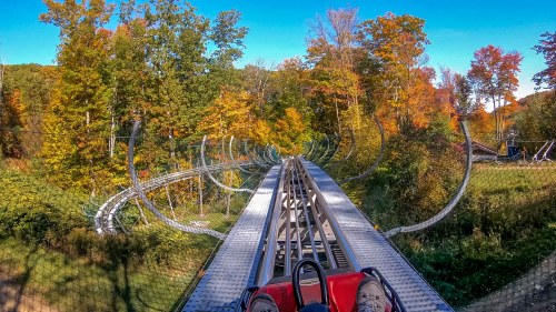 A view from a roller coaster track winding through colorful autumn trees under a clear blue sky.