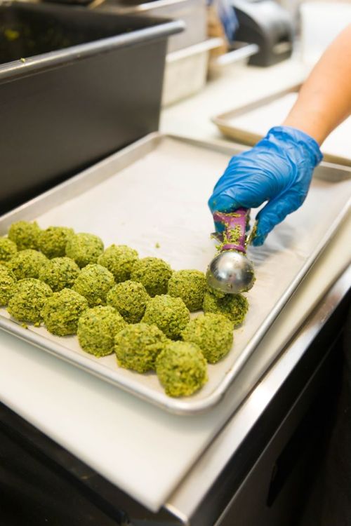 A hand in a blue glove scoops green mixture into round balls on a baking tray.