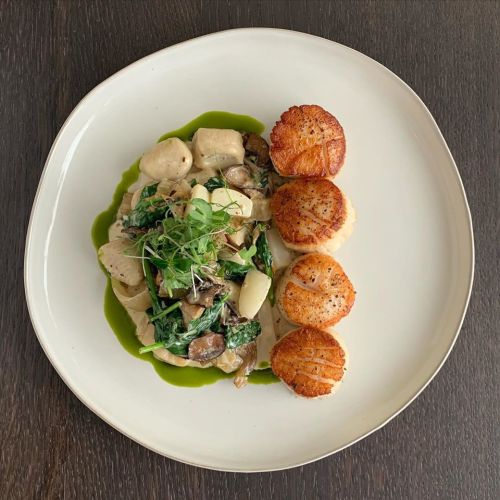 A plate of gnocchi with mushrooms and greens, topped with three seared scallops and a drizzle of green sauce.