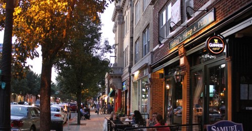 A lively street scene at dusk, featuring shops, outdoor seating, and trees with warm lighting.