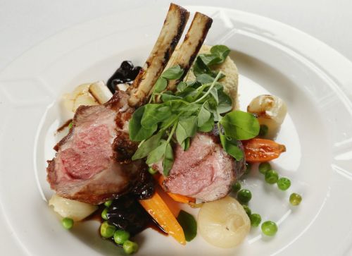 A gourmet dish featuring two lamb chops, colorful vegetables, and a garnish of fresh greens on a white plate.