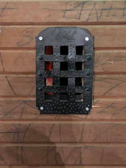 A decorative black metal vent cover with a grid pattern mounted on a wooden wall.