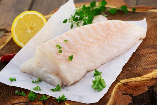 Fresh fish fillet on parchment paper, garnished with herbs and a slice of lemon, on a wooden cutting board.