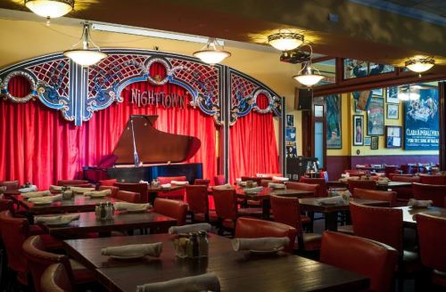 A cozy restaurant interior featuring red curtains, a grand piano on stage, and neatly arranged tables.