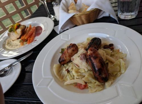 A plate of grilled sausages on a bed of cabbage and tomatoes, with a side of bread and a glass of water in the background.
