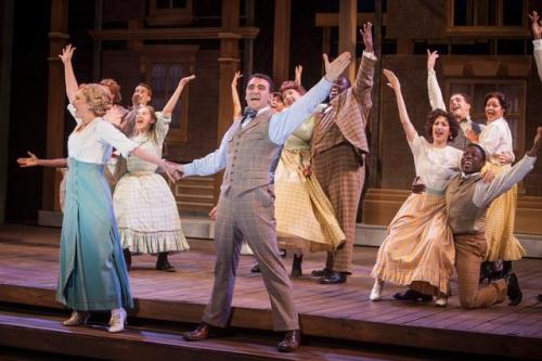 A lively musical performance with a diverse cast, featuring dancers in period costumes celebrating on stage.