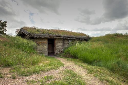 A rustic wooden hut with a grass-covered roof, surrounded by tall green grass and a cloudy sky.