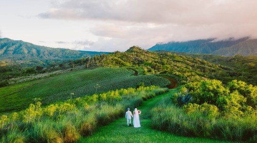 A couple walks hand in hand along a lush green path, surrounded by rolling hills and mountains under a cloudy sky.