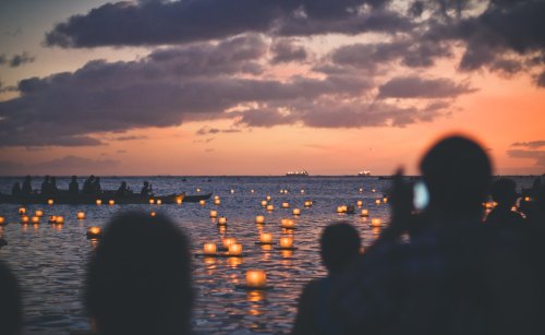 People gather by the water at sunset, releasing glowing lanterns into the calm sea.