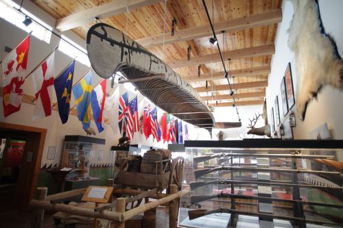 Interior of a museum featuring a large canoe hanging from the ceiling, surrounded by flags and historical artifacts.