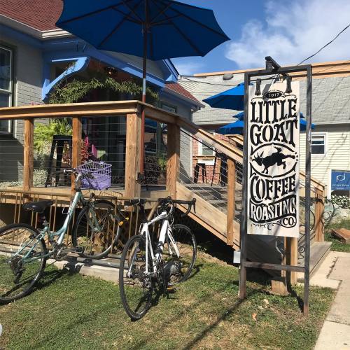 A cozy coffee shop with a wooden deck, blue umbrellas, and bicycles parked outside. Sign reads "Little Goat Coffee Roasting Co."
