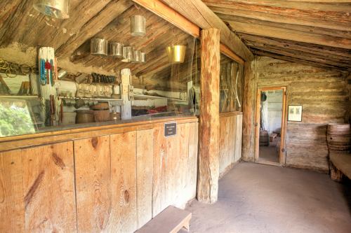 Interior of a rustic wooden building with shelves, tools, and a door, showcasing a historical setting.
